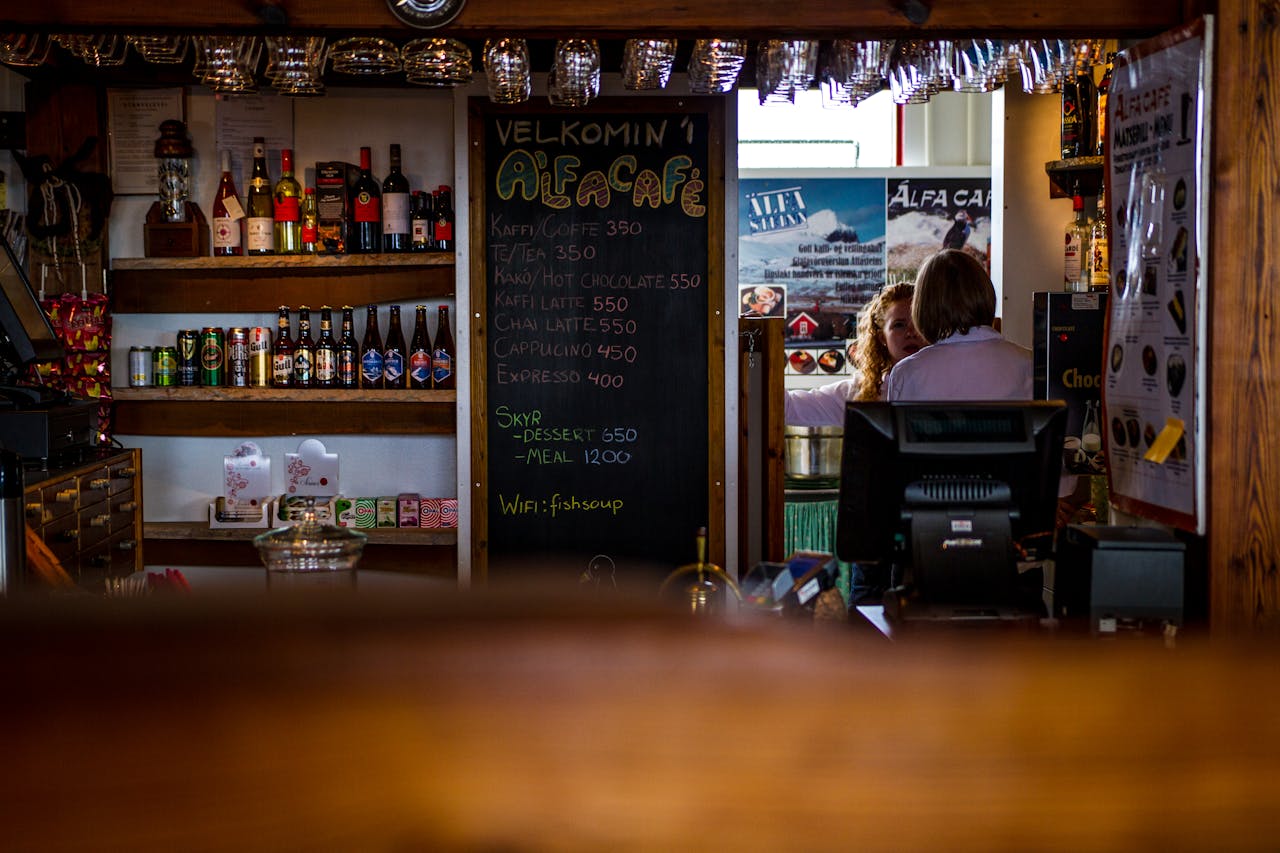 Warm and inviting café scene in Iceland featuring a menu chalkboard, drinks on shelves, and people in conversation.